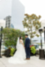 Bride in white gown and groom in blue suit holding hands, standing on a terrace with greenery and skyscraper backdrop. Overcast day at Terminal City Club Wedding