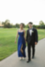 Couple walking hand in hand on a pathway, woman in blue dress, man in black suit, greenery in background, evening setting, smiling at Mayfair Golf Course Wedding
