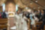 Bride and groom in formal attire walk down the aisle of a church, smiling, with guests applauding. Stained glass and wooden pews visible/St. John the Apostle Parish wedding ceremony Vancouver