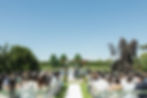 Outdoor wedding ceremony, couple stands under floral arch. Guests seated on white chairs. Sunny day with blue sky and lush greenery around at Mayfair Golf Course Wedding