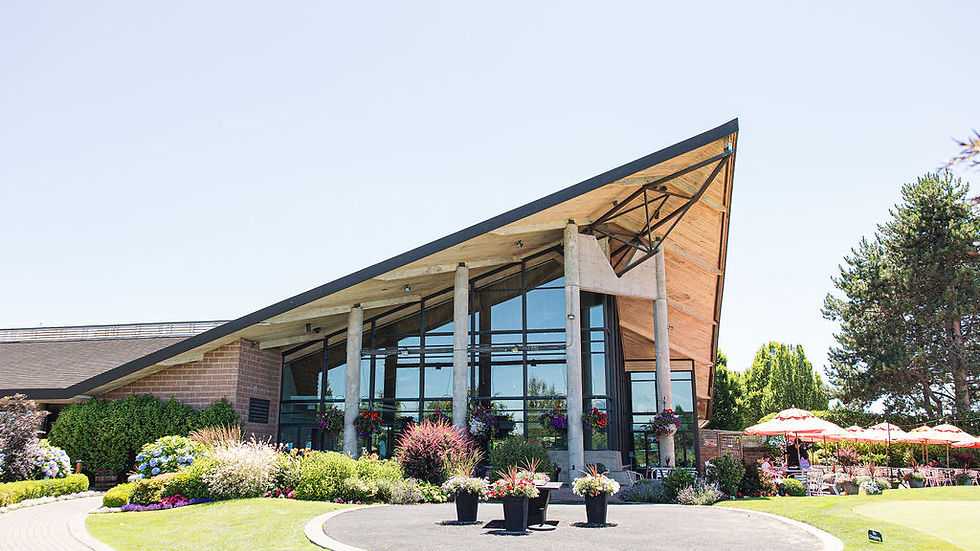 Modern building with a slanted roof, surrounded by vibrant gardens and trees. Red umbrellas cover tables to the right under a clear sky /Mayfair Golf Course Wedding