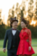 A couple in formal attire with playful hats walks in a park at sunset, smiling. The woman wears a red dress; the background is blurry /Quilchena Golf Club wedding photos Richmond