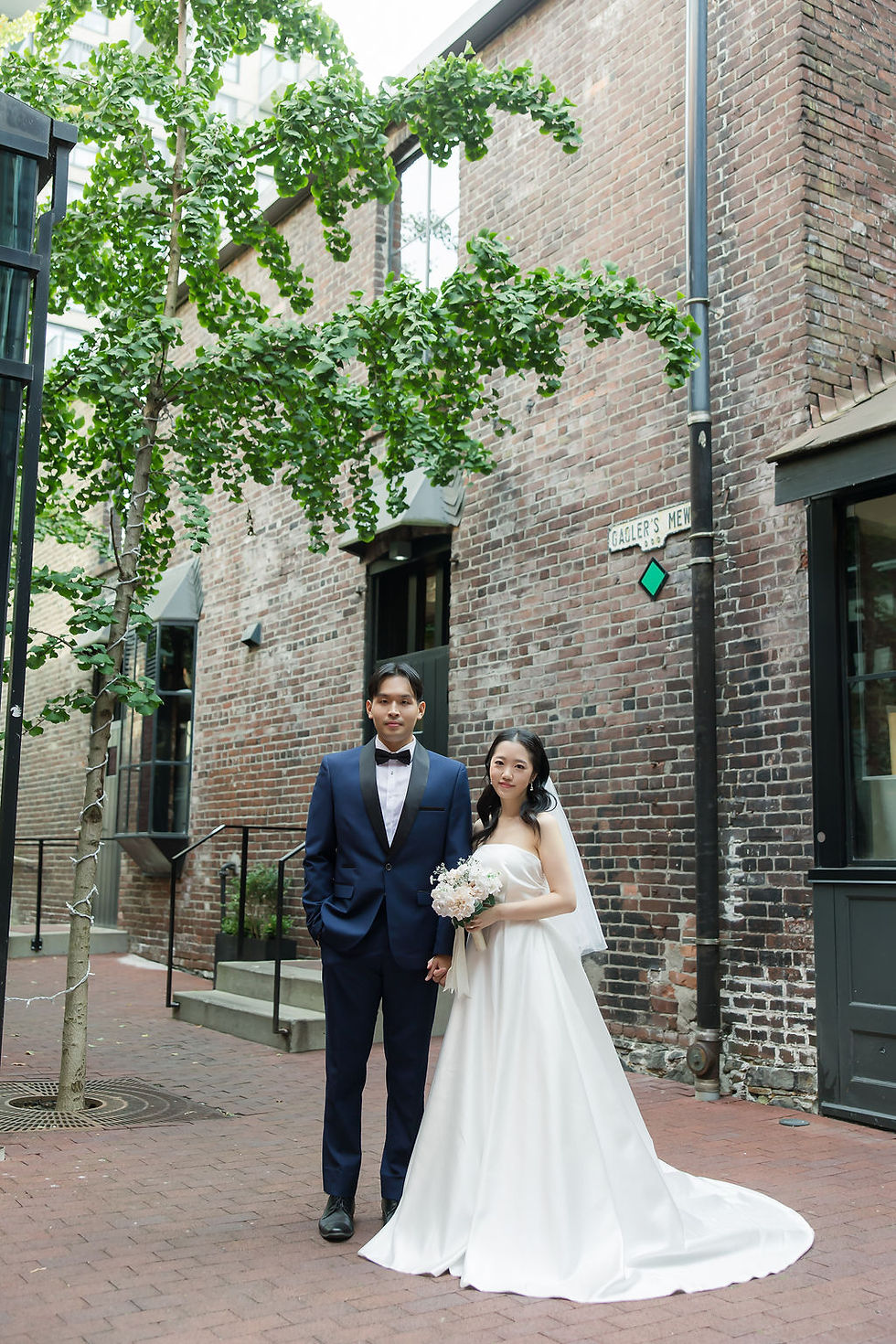 Bride in white gown and groom in navy suit pose on a brick street. The bride holds a bouquet. Background features a tree and brick walls and Pre Wedding Photoshoot Vancouver – One-Stop Gastown Experience with The Eunice & Co.
