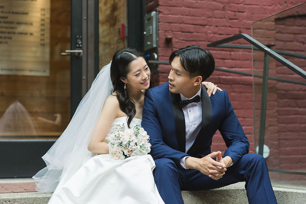 Bride in white dress and veil sits with groom in blue suit on steps. They gaze at each other affectionately. Red brick backdrop. Pre Wedding Photoshoot Vancouver – One-Stop Gastown Experience with The Eunice & Co.