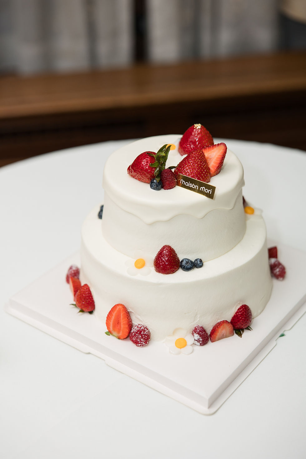 Two-tier white cake with strawberries, blueberries, and raspberries. "Maison Mori" label on top. Simple white table setting at The Vancouver Club