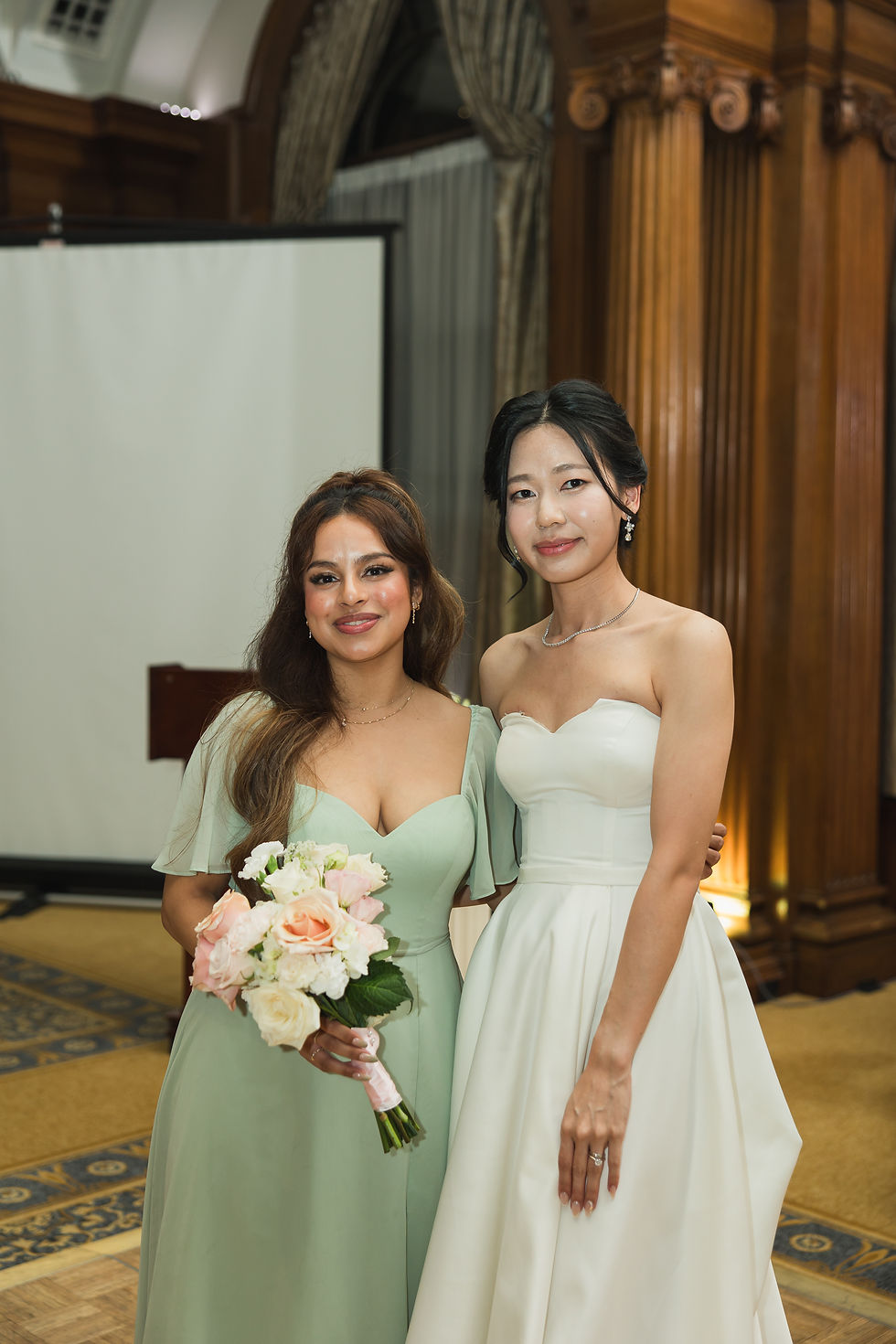 Two women in formal dresses pose in a decorated room; one in mint holding flowers, the other in white, both smiling warmly. Taking photo after bouquet toss in The Vancouver Club