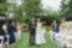 Bride in white dress and man in suit walk down outdoor aisle at garden wedding. Guests clap and take photos. Lush greenery and flowers at VanDusen Botanical Garden Wedding and Shaughnessy Restaurant wedding