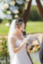 Bride in white dress reads vows with a microphone outdoors, under floral arch. Bright, sunny day with greenery in the background at Mayfair Golf Course Wedding