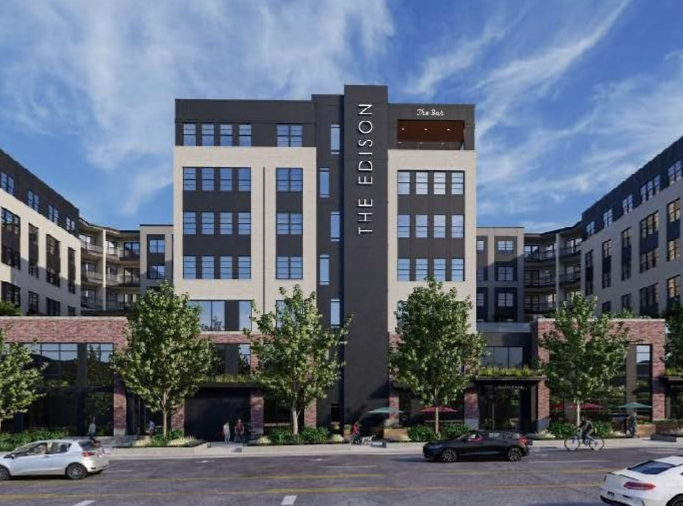 Modern building with "The Edison" sign, surrounded by trees. Cars and cyclists on street. Clear blue sky, several people walking.