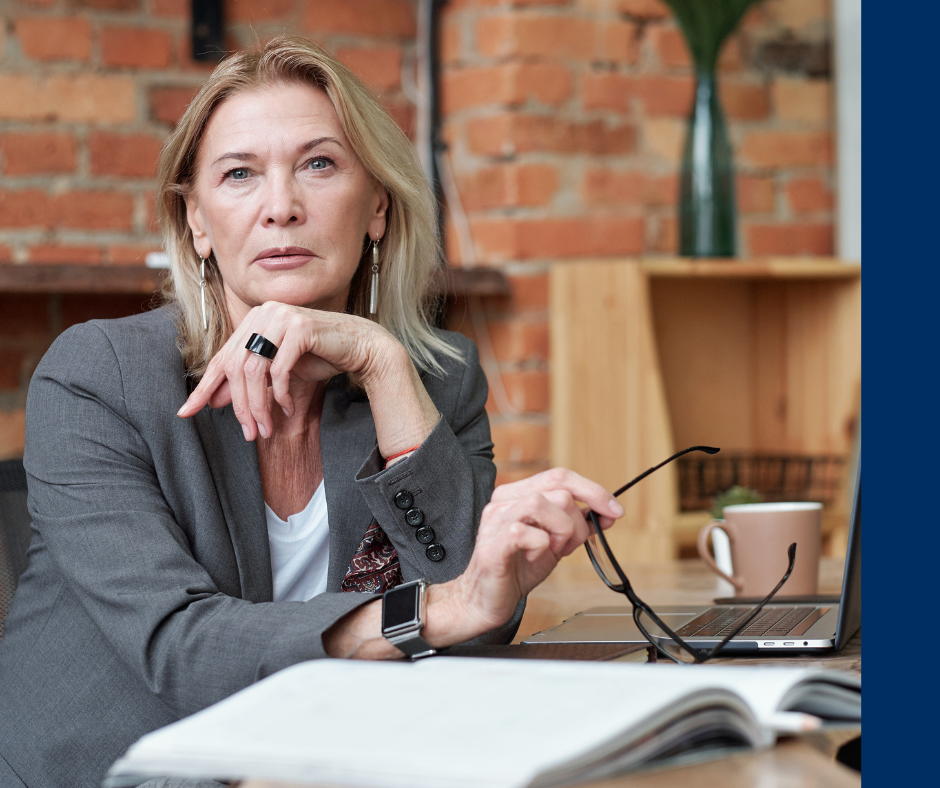 Woman in a gray suit holds glasses, looking thoughtful. Brick wall and laptop in background, with open notebook in foreground. Calm mood.