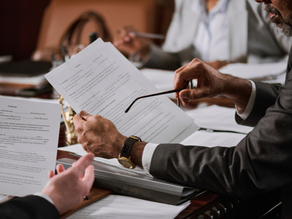 Meeting with people in suits reviewing contracts at a wooden table. Papers, pens, and documents are scattered. Focused, professional mood.