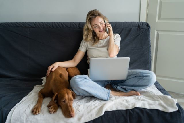 girl talking on cell phone with computer on her lap and arm resting on her dog by her side on large dark blue futon - in home pet euthanasia near me