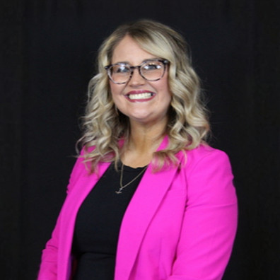 Smiling woman with blonde wavy hair and glasses wearing a bright pink blazer over a black top, posed against a dark background