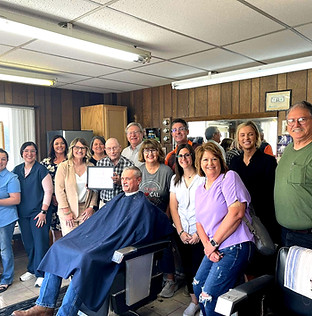 Group of community members gathered inside a barbershop, smiling around a seated man in a barber chair, suggesting a business of the month