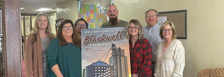 Chamber members standing beside an easel displaying a community guide poster that reads “Welcome Home to Blackwell,” featuring a sunset scene with a grain elevator painted with an American flag