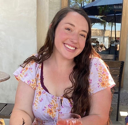 a woman with long dark brown wavy hair with brown eyes. She is wearing a pink floral dress, has a nose ring and is smiling. You can see a cafe with chairs and tables in the backgrounds. There is a pink beverage in front of her. 