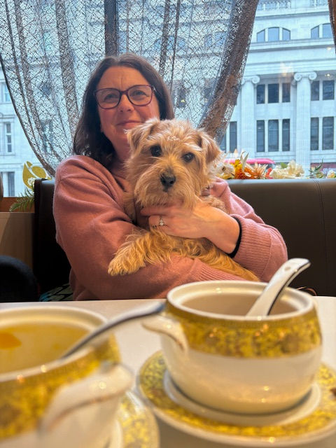 Woman in glasses holds a dog at a cafe table with two ornate cups. Large window with city view in background; relaxed, cozy mood.