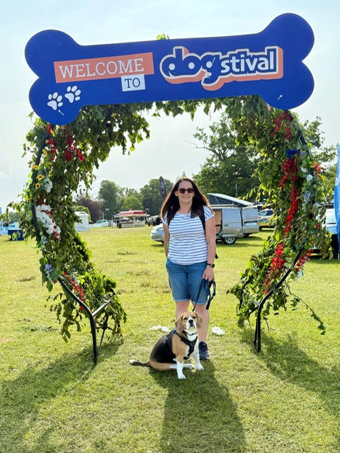 Woman with dog under a Welcome to dogstival bone-shaped arch at a sunny outdoor festival.