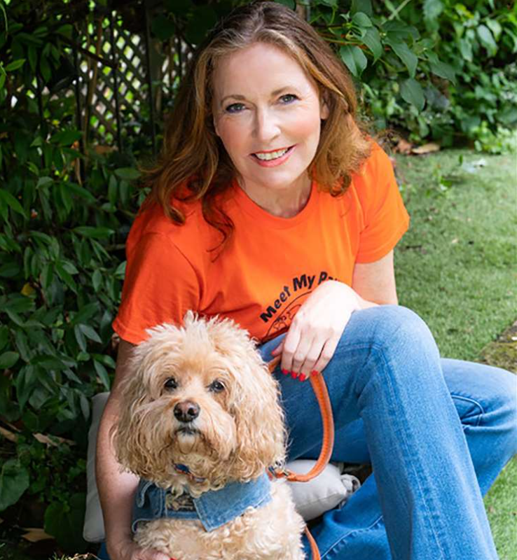 Smiling woman in an orange shirt with "Meet My Pet" text, kneels beside a fluffy dog in a garden. The dog wears a denim vest.