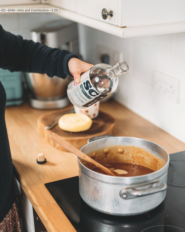   Hand pours clear liquid from a bottle into a pot of sauce on a kitchen counter, with a wooden spoon and cheese wheel nearby.