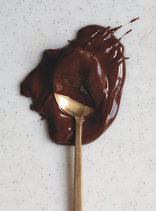 Gold spoon resting in a glossy dollop of melted chocolate on a white speckled surface.