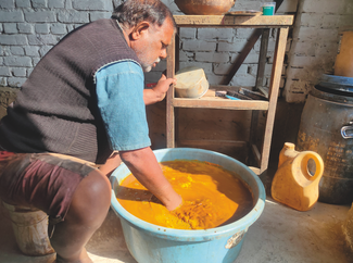 Man kneels in a workshop, stirring orange liquid in a large blue tub beside shelves and containers in warm sunlight.