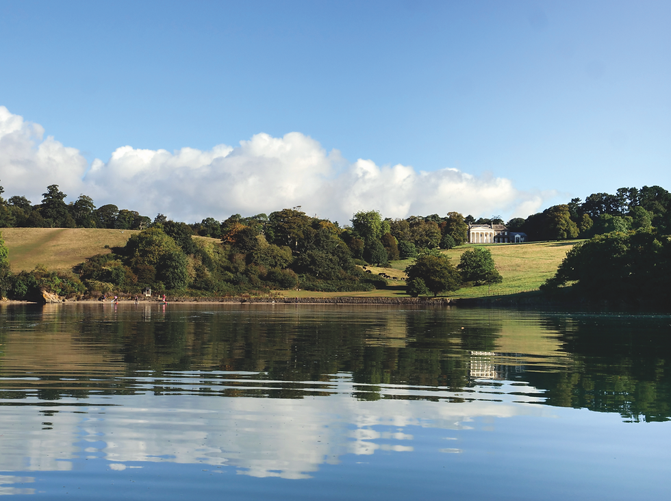 Calm lake reflects blue sky and clouds, with green hills, trees, a white mansion, and tiny paddleboarders in the distance.