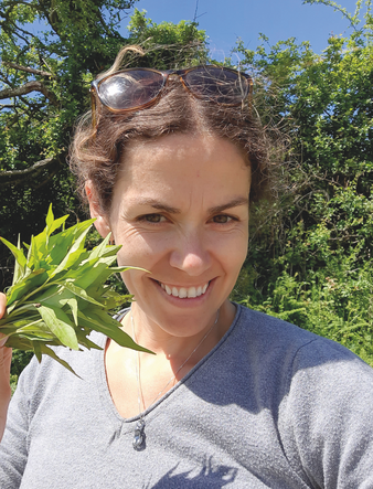 Smiling woman outdoors holds a bunch of green leaves, sunglasses on head, with sunlit trees and blue sky behind her.