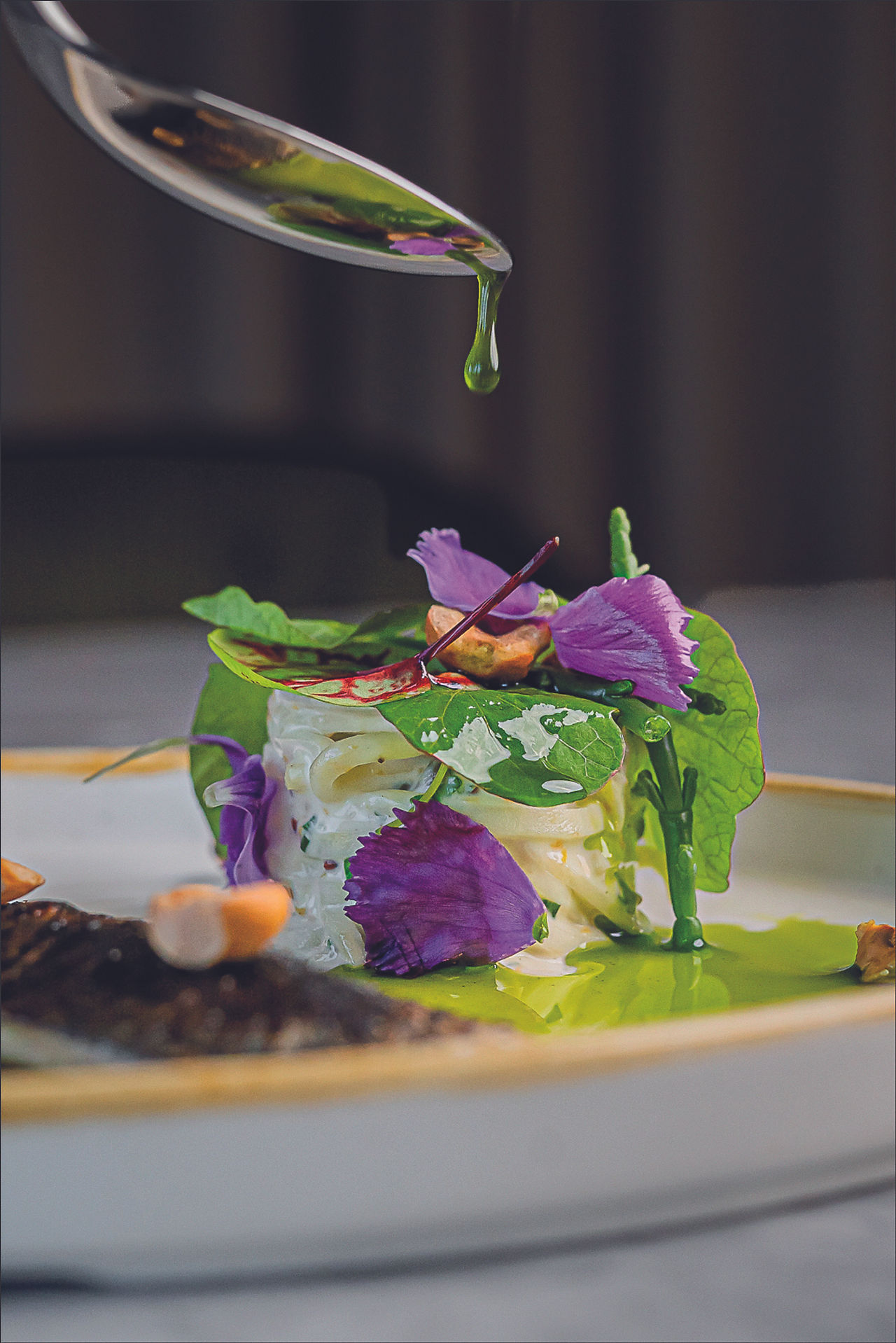 Spoon dripping green oil over a gourmet dish garnished with purple flowers and greens on a white plate