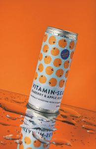 Tilted Vitamin Sea seaberry and apple juice can on bright orange background, with water splashes and a mirrored reflection.