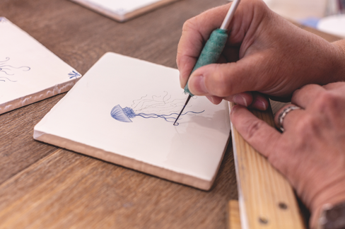 Close-up of a hand drawing a blue jellyfish on a white tile with a fine tool at a wooden table.