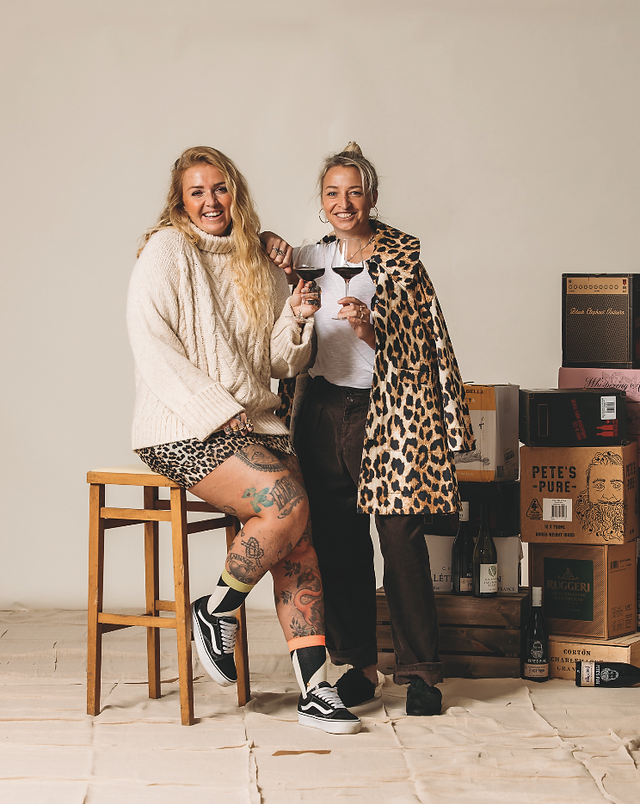 Smiling two women hold wine glasses beside stacked wine boxes; one sits on a stool in a cream sweater, the other wears leopard coat.