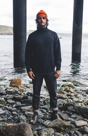 Man in orange beanie and black sweater stands on rocky shore beneath pier pillars by calm water. Finisterre