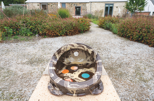 Decorative ceramic bowl sculpture on a pedestal in a gravel garden path, with a stone building and shrubs in the background.