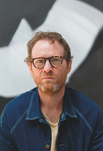 Artist Andrew Bryant. Portrait of a serious man with glasses and a denim jacket against a blurred black-and-white background.
