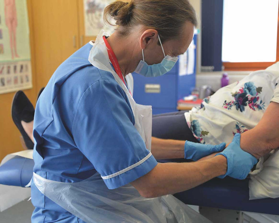 Masked nurse in blue scrubs and gloves helps a patient on an exam table in a clinic room.