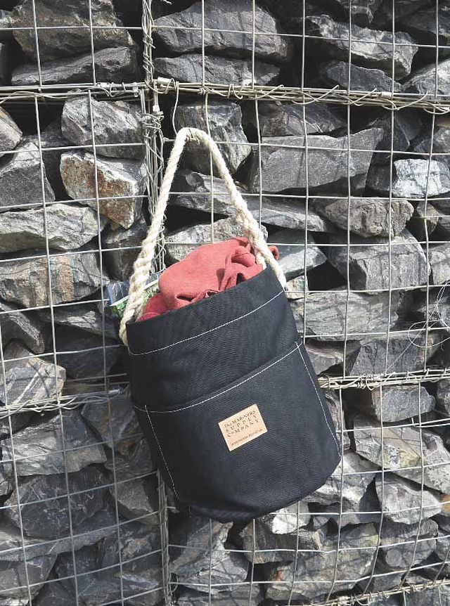 Black rope-handled tote with red cloth inside hangs on a wire rock wall, with a small tan label.