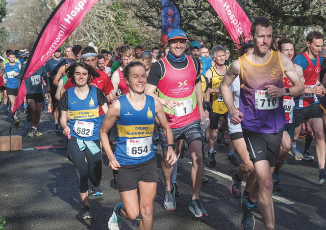Runners in a sunny road race pass pink Cornwall Hospice banners, with bib numbers and club jerseys, smiling and focused.