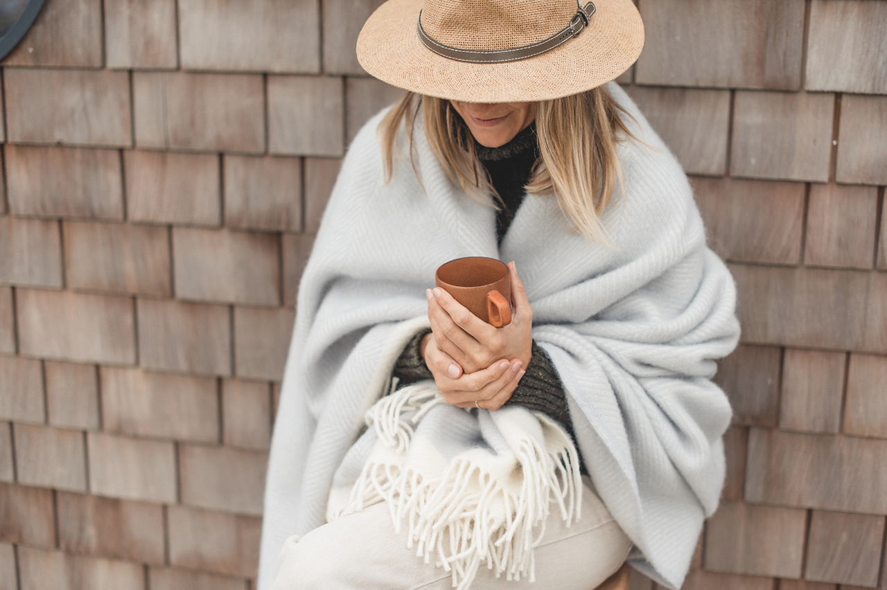 Woman in a straw hat sits wrapped in a gray blanket, holding a brown mug against a weathered shingle wall. Blankets woven by Atlantic Blankets