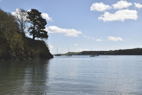 National Trust Cornwall. Calm bay with rocky tree-lined cliff, anchored sailboats, and a bright blue sky with scattered clouds.