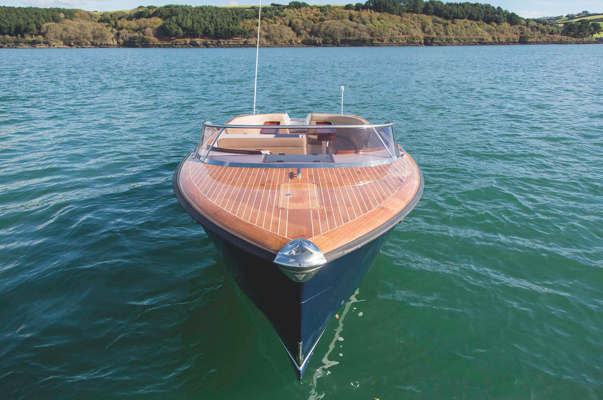 Luxury wooden motorboat on calm blue water, facing camera, with wooded shoreline in the background under a clear sky