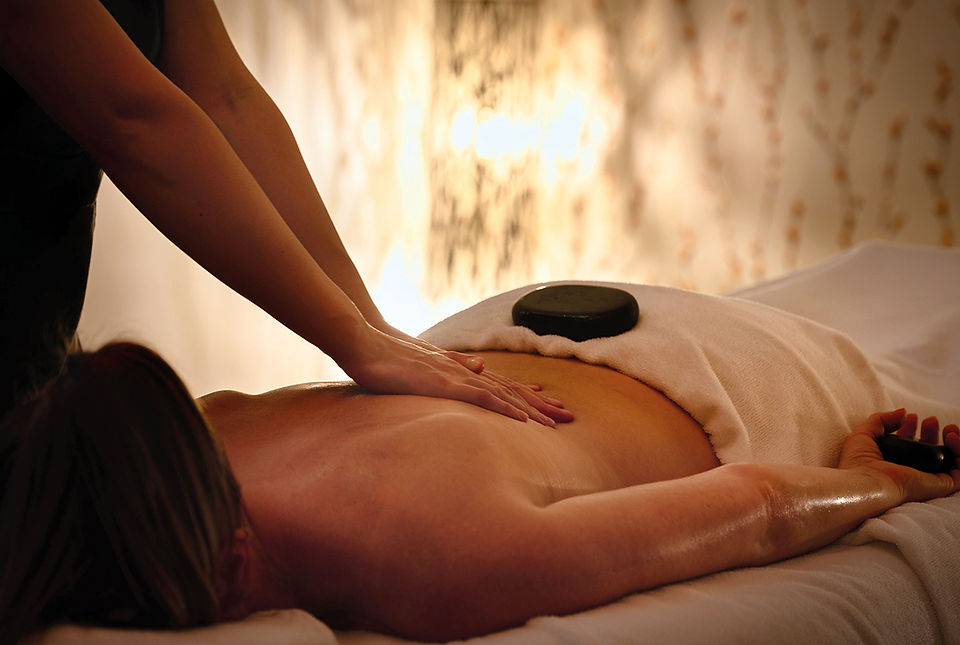 Spa therapist massages a client’s back with hot stones on a massage table in warm, calming light.
