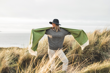 Woman in a black hat holds a green shawl open in grassy dunes by the sea, looking calm and content. Blankets woven by Atlantic Blankets