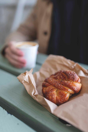 A person holds a coffee cup beside a sugar-dusted braided pastry on a tray, with a soft blurred cafe background.