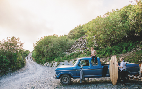 Shirtless man sits on a blue pickup while another holds a surfboard beside it on a rocky rural road with walkers in the distance.