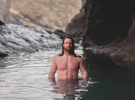 Anthony mullally Shirtless bearded man stands waist-deep in a rocky sea cave, looking serious in calm water.