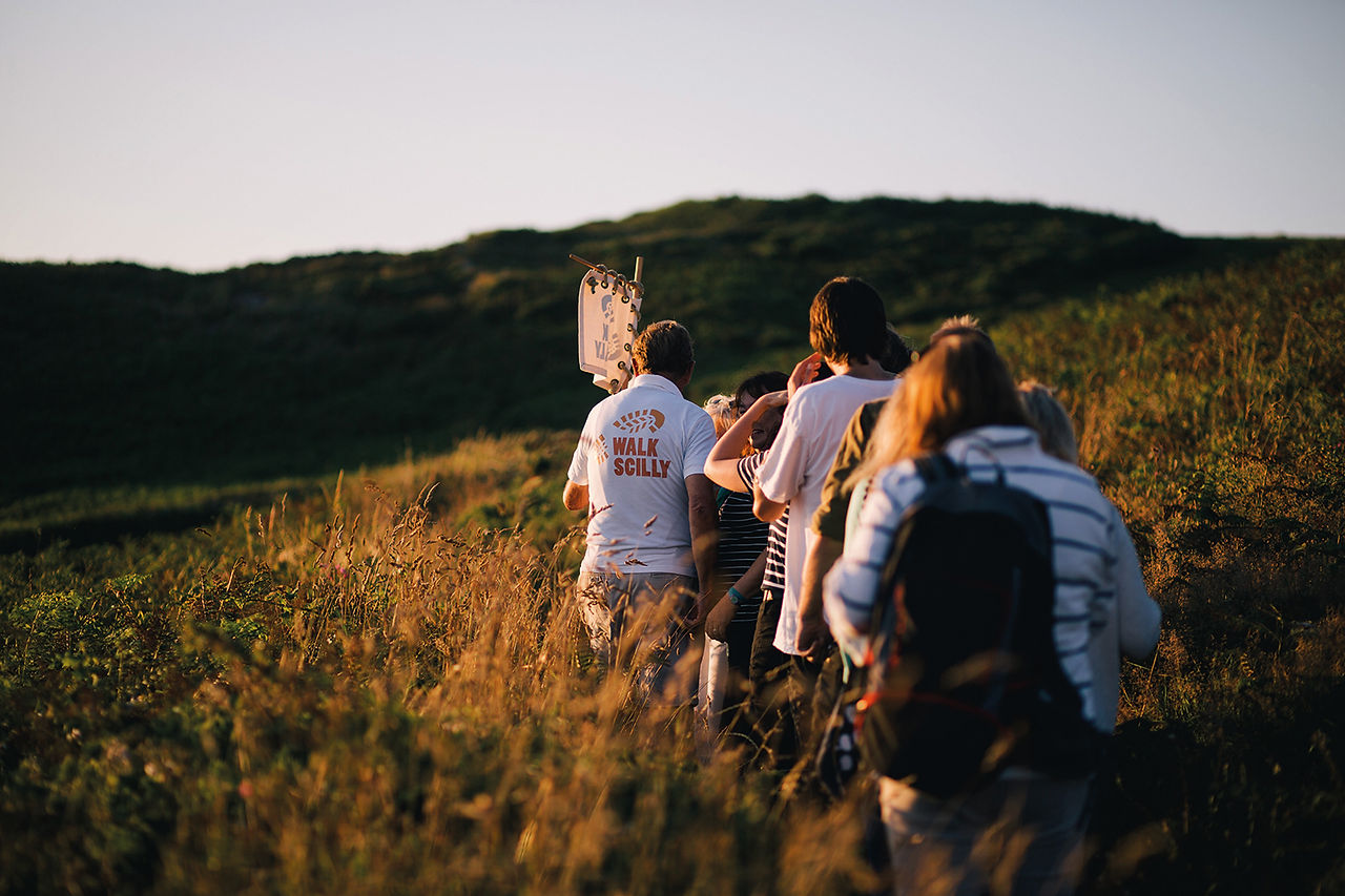 Group hiking through sunlit grassy dunes, one shirt reads WALK SCILLY.