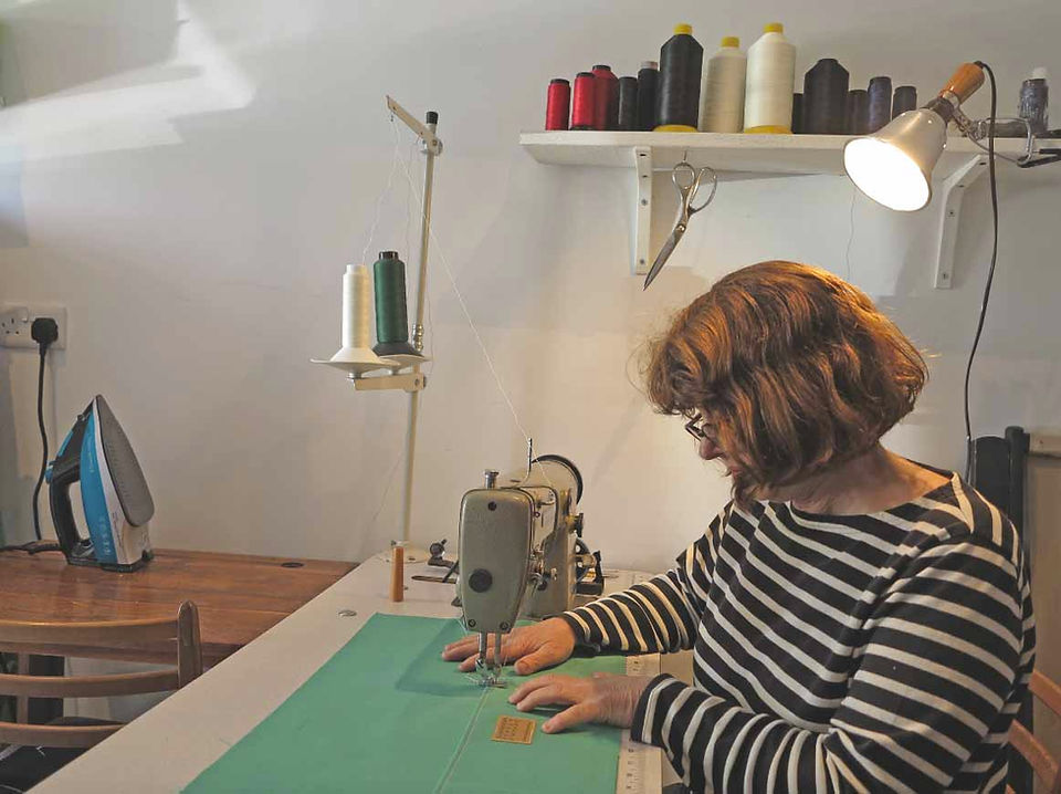  Woman in striped shirt sews green fabric on a machine in a bright sewing room, with thread spools, scissors, and an iron nearby.