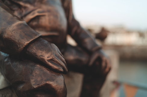 Close-up of a bronze statue’s hands resting on a knee, with a softly blurred outdoor background and warm, reflective light.