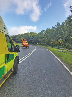 Ambulance van beside a red rescue helicopter on a curving wet road, with chevron signs, trees, and blue sky.